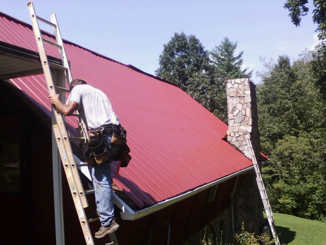 Roofer on a ladder beside a red metal roof and stone chimney — Soffit & Fascia Repair by Roofing Brothers in Bridger